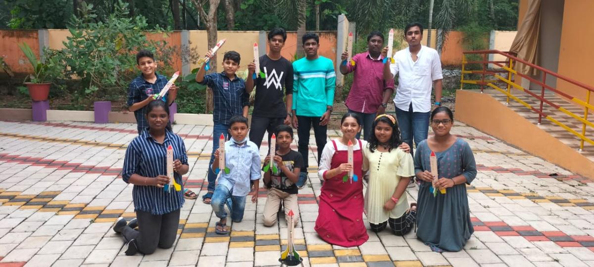 Students and mentors gathered outdoors holding colourful model rockets, with a larger rocket set upright in front of the group