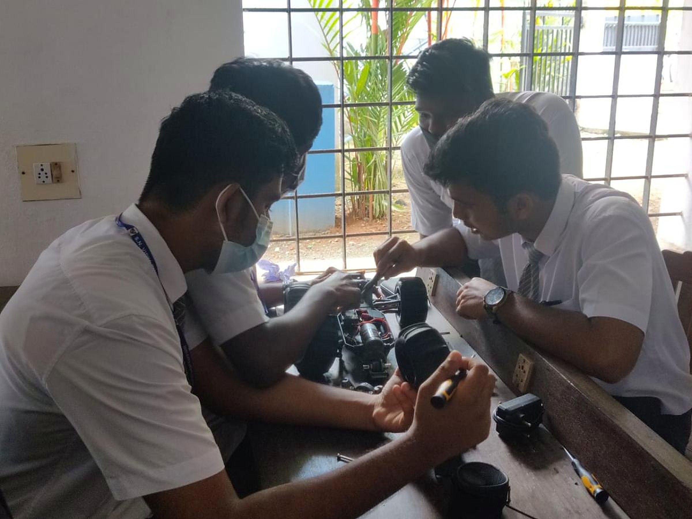 Four students in school uniform leaning over a workbench with screwdrivers and tools, stripping down a motor and electronics assembly