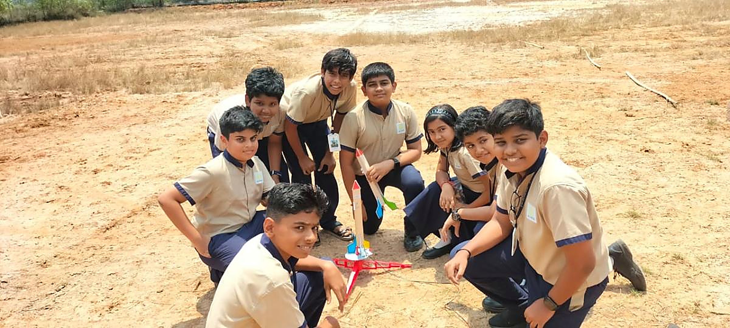 A group of school students crouched around a model rocket on its launch pad on an open dirt field, moments before launch