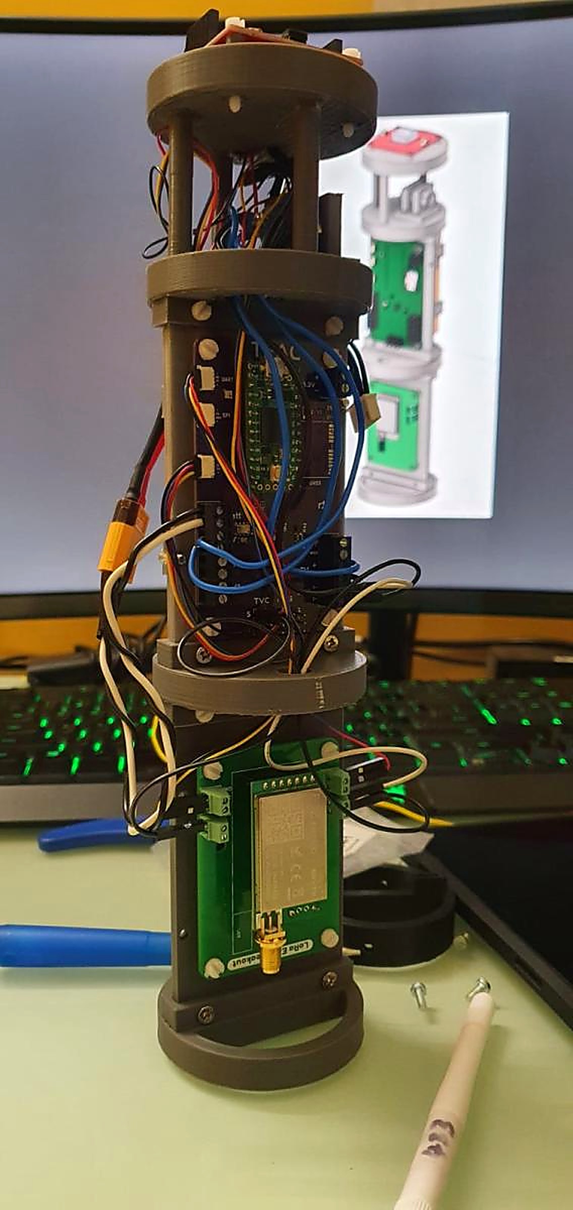 A 3D-printed rocket avionics bay with stacked circuit boards, GPS module and coloured wiring, with its CAD model displayed on the monitor behind