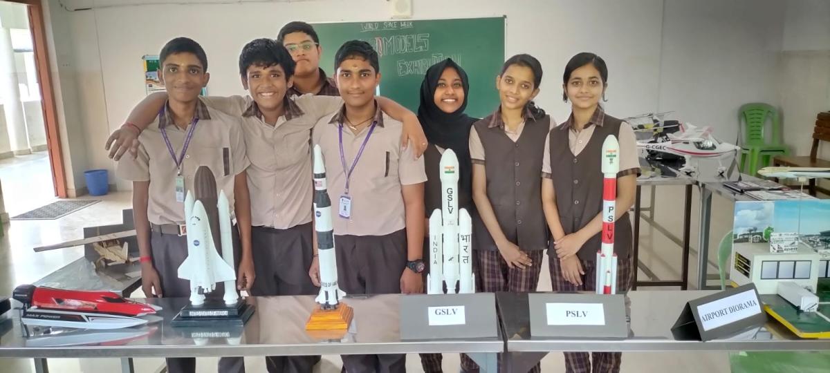 Seven school students standing behind a display of space shuttle, GSLV and PSLV models alongside an airport diorama