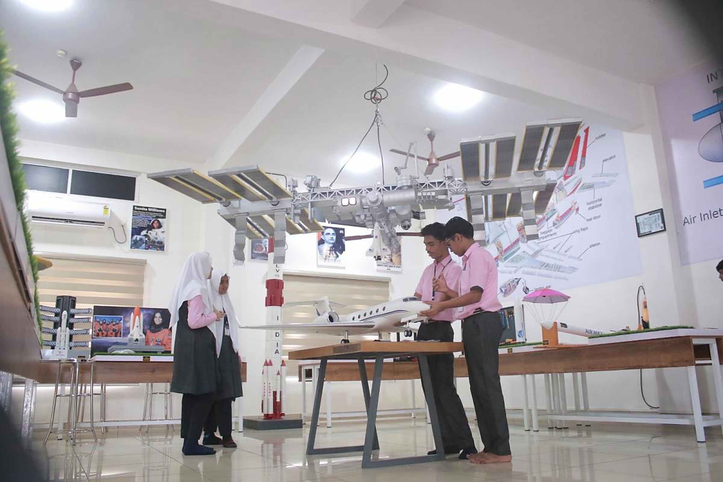 Students examining a business jet model in an aerospace lab hung with a large space station model, launch vehicle models and aerodynamics posters